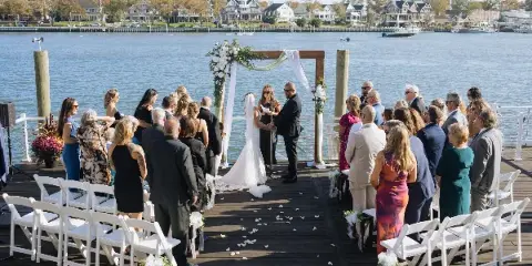 A wedding ceremony taking place on a dock with a wooden arch, white chairs, and floral arrangements.
