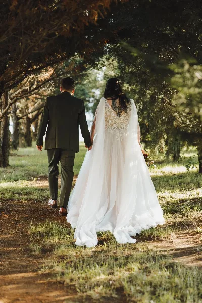 A bride and groom walking hand-in-hand down a tree-lined path outdoors.