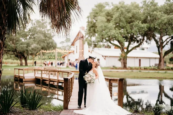 A bride and groom kissing on a wooden bridge over a pond with a white barn and oak trees in the background.