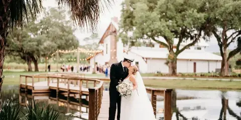 A bride and groom kissing on a wooden bridge over a pond with a white barn and oak trees in the background.
