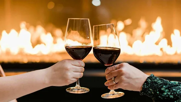 Two hands clinking glasses of red wine in a toast with a fireplace in the background.