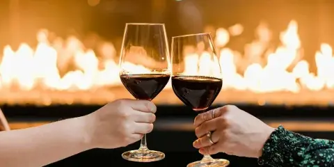 Two hands clinking glasses of red wine in a toast with a fireplace in the background.
