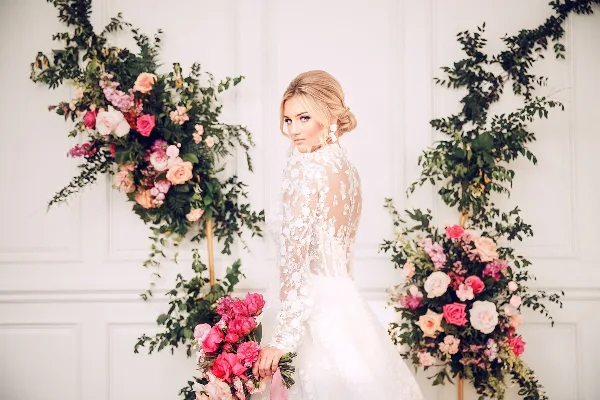 Bride in a lace wedding gown holding a pink bouquet beside floral arrangements against a white paneled wall.