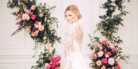 Bride in a lace wedding gown holding a pink bouquet beside floral arrangements against a white paneled wall.