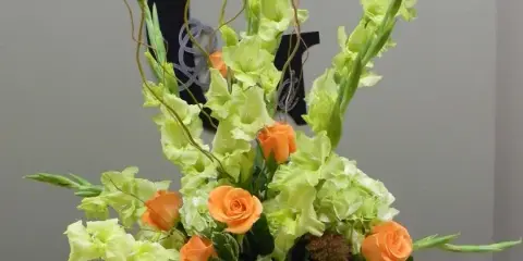 A tall floral arrangement with orange roses and lime-green gladiolus in a square vase