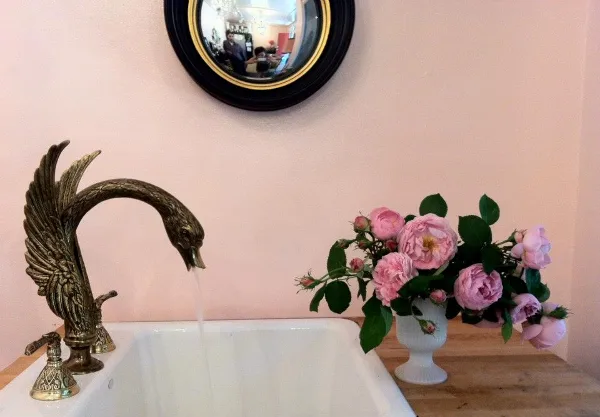 A floral arrangement of pink roses in a white vase placed on a wooden counter next to a decorative bird-shaped faucet.