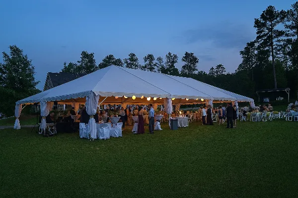 A large white event tent set up on a grassy lawn at dusk, filled with tables, chairs, and guests.