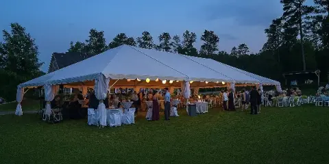 A large white event tent set up on a grassy lawn at dusk, filled with tables, chairs, and guests.