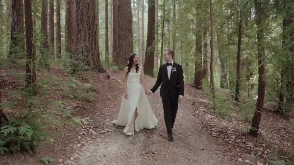 A bride and groom holding hands and walking down a path in a forest.