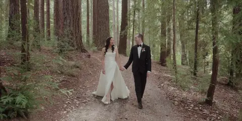A bride and groom holding hands and walking down a path in a forest.