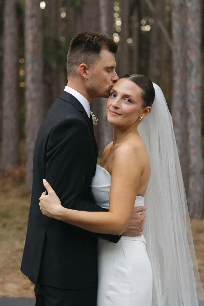 A groom in a black suit kisses the forehead of a bride in a white dress and veil, standing in a forest setting.