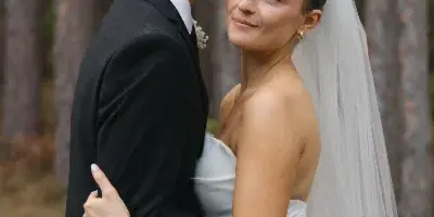 A groom in a black suit kisses the forehead of a bride in a white dress and veil, standing in a forest setting.