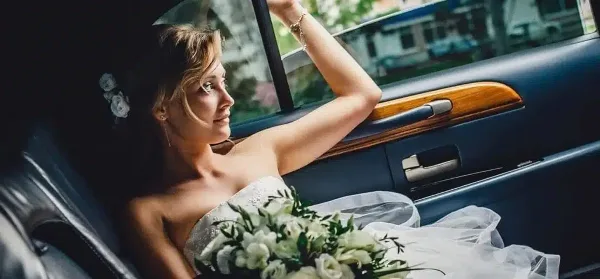 A bride in a white wedding dress and holding a bouquet sits in the back seat of a luxury car.