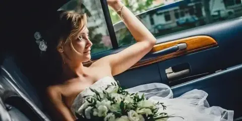 A bride in a white wedding dress and holding a bouquet sits in the back seat of a luxury car.