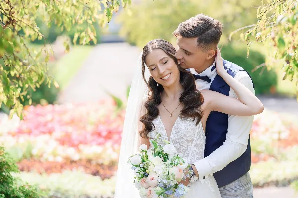 A bride and groom posing together outdoors, with the groom kissing the bride's cheek.