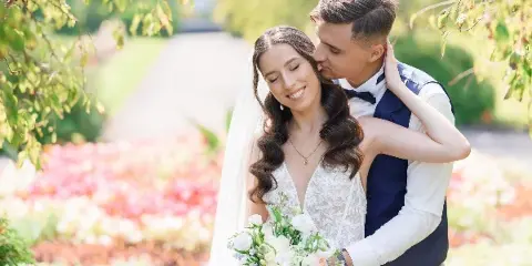 A bride and groom posing together outdoors, with the groom kissing the bride's cheek.