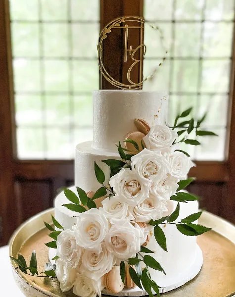 A three-tier white wedding cake decorated with cascading white roses, greenery, and macarons, topped with a gold monogram topper.