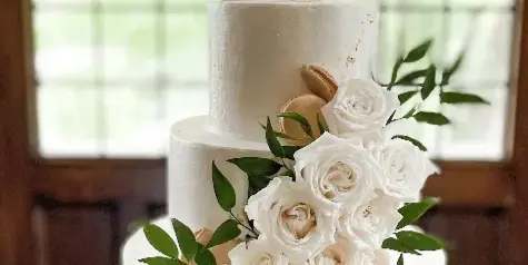 A three-tier white wedding cake decorated with cascading white roses, greenery, and macarons, topped with a gold monogram topper.