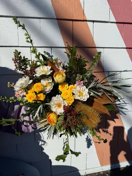 A close-up of a vibrant floral arrangement with yellow roses, white ranunculus, and greenery held against a striped wall.