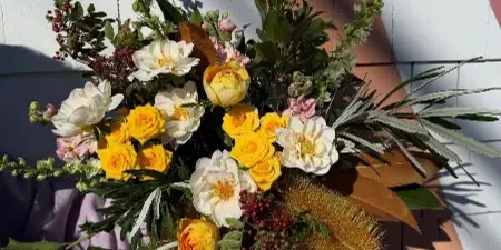 A close-up of a vibrant floral arrangement with yellow roses, white ranunculus, and greenery held against a striped wall.