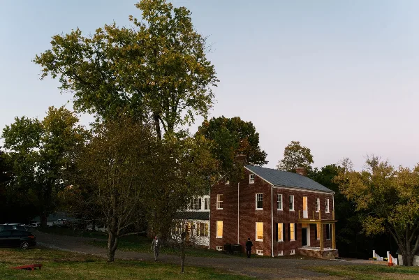 Exterior view of a historic two-story red brick estate building with white trim, surrounded by mature trees and a grassy lawn at dusk.