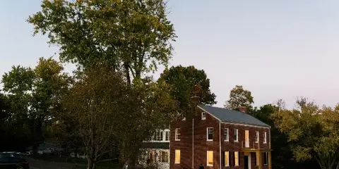 Exterior view of a historic two-story red brick estate building with white trim, surrounded by mature trees and a grassy lawn at dusk.