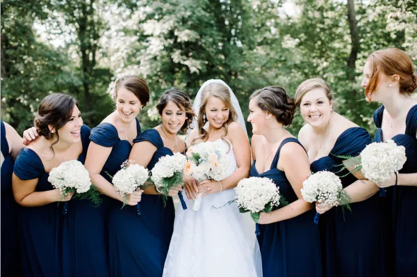 A bride in a white gown and veil stands smiling with a group of bridesmaids in navy blue dresses, all holding white bouquets outdoors.