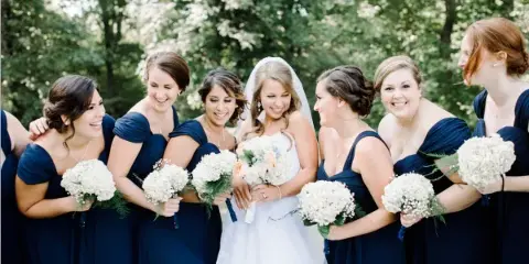 A bride in a white gown and veil stands smiling with a group of bridesmaids in navy blue dresses, all holding white bouquets outdoors.