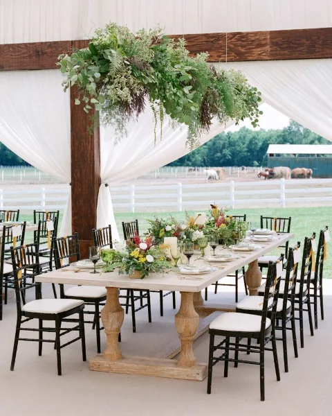 Luxury tented farm wedding reception featuring a long wooden table, black chiavari chairs, and lush floral centerpieces under a white tent.