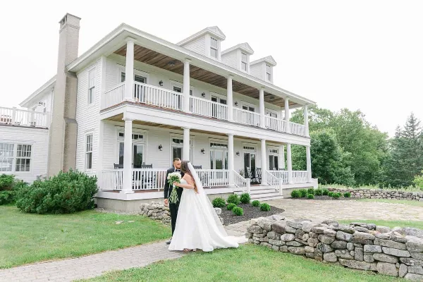 A bride and groom standing on a stone path in front of a large white two-story manor house with a porch.