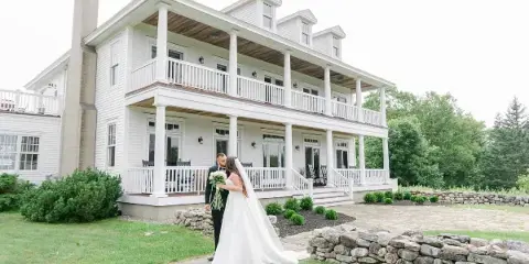 A bride and groom standing on a stone path in front of a large white two-story manor house with a porch.