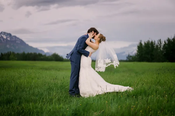 A bride and groom kissing in a green field with mountains in the background.