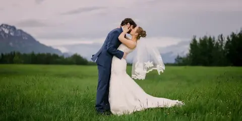 A bride and groom kissing in a green field with mountains in the background.