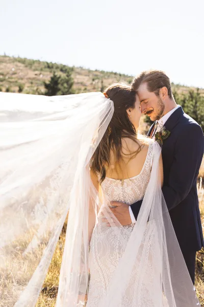 A bride and groom embracing in a field with the bride's veil blowing in the wind.