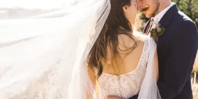 A bride and groom embracing in a field with the bride's veil blowing in the wind.