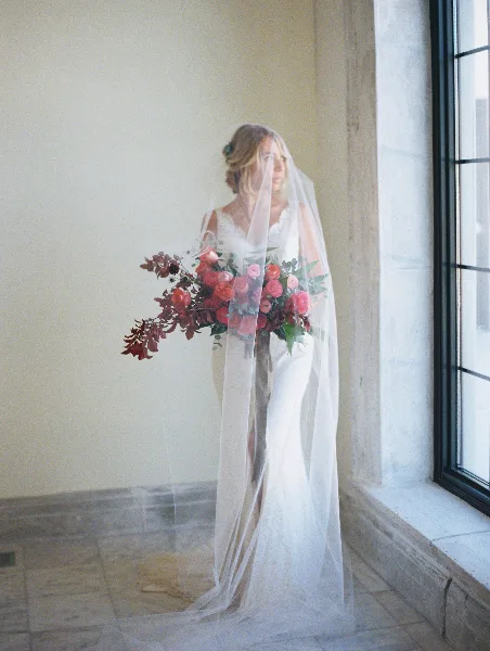 A bride in a white gown and veil holding a large, organic bouquet of red and pink roses with dark foliage.