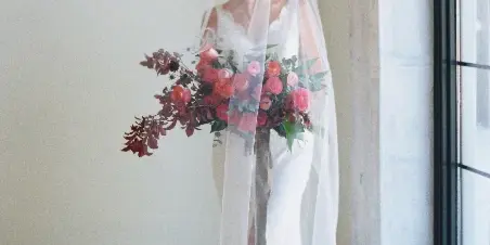A bride in a white gown and veil holding a large, organic bouquet of red and pink roses with dark foliage.