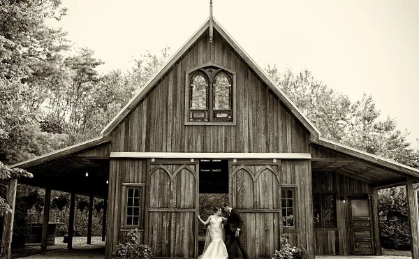 A black and white photograph of a couple standing in front of a rustic wooden barn with Gothic-style windows and doors.