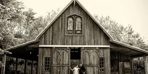 A black and white photograph of a couple standing in front of a rustic wooden barn with Gothic-style windows and doors.