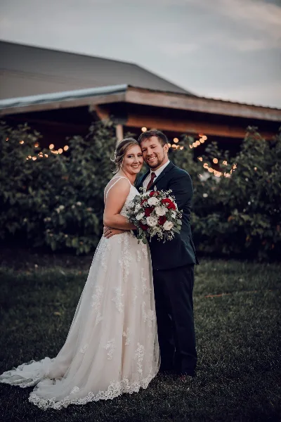 A bride and groom standing in front of a rustic barn with string lights and greenery.