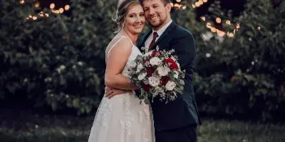 A bride and groom standing in front of a rustic barn with string lights and greenery.