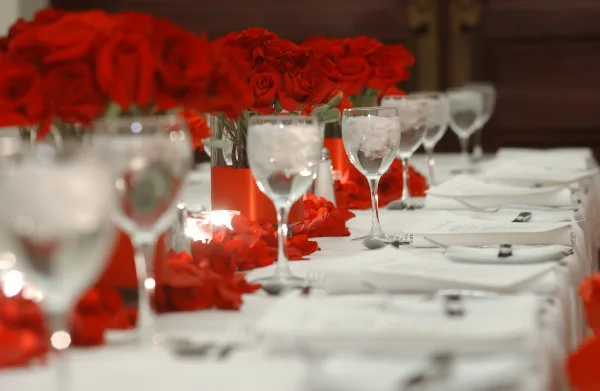 A close-up view of a formal event table setting with red rose centerpieces, wine glasses, and white linens.