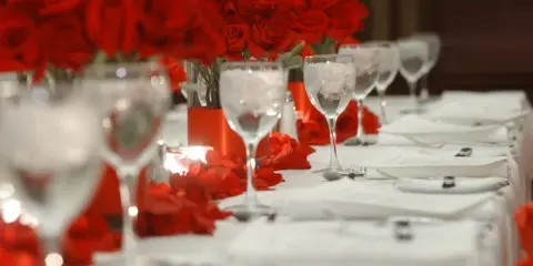 A close-up view of a formal event table setting with red rose centerpieces, wine glasses, and white linens.