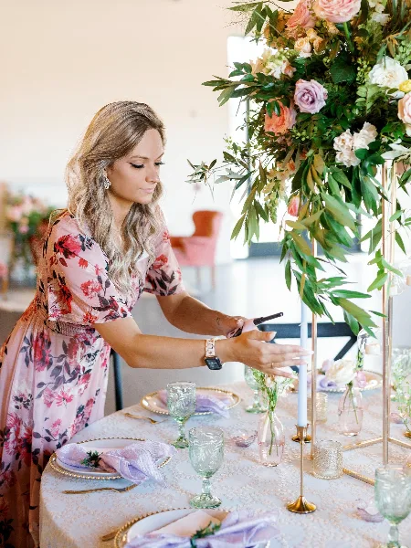 A woman in a floral dress arranging a wedding table setting with tall floral centerpieces and gold candlesticks.