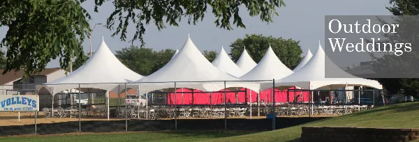 A row of white peak tents set up for an outdoor wedding event with tables and chairs arranged underneath.