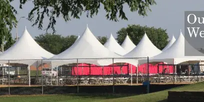 A row of white peak tents set up for an outdoor wedding event with tables and chairs arranged underneath.