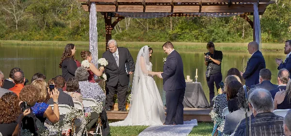 A bride and groom exchanging rings during an outdoor wedding ceremony on a wooden stage next to a pond, with guests seated in the foreground.