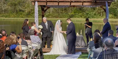 A bride and groom exchanging rings during an outdoor wedding ceremony on a wooden stage next to a pond, with guests seated in the foreground.