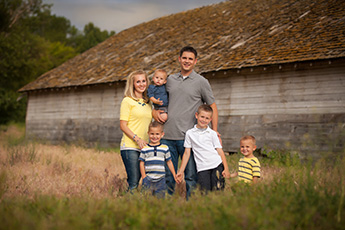 A family of six posing for a portrait outdoors in front of a rustic wooden barn.