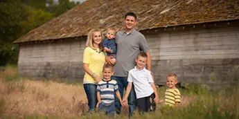 A family of six posing for a portrait outdoors in front of a rustic wooden barn.